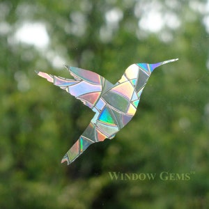 May include: A iridescent hummingbird window cling with a stained glass design. The cling is made of a durable vinyl material and is easy to apply and remove. The hummingbird is facing right and has its wings spread out. The background is a blurry green.