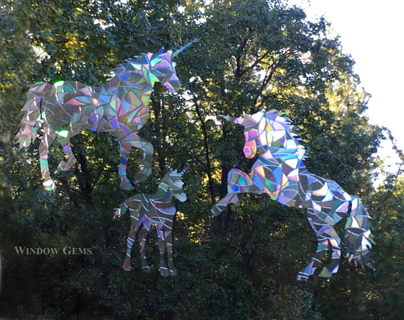 May include: Three iridescent unicorn window clings, one large, one medium, and one small, against a green leafy background. The unicorns are made of iridescent mosaic pieces.