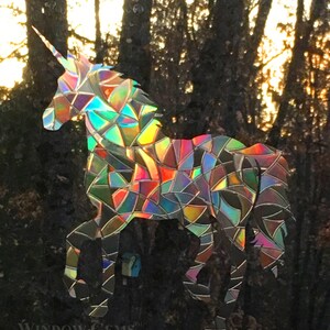 May include: A rainbow iridescent unicorn silhouette window cling. The unicorn is made of a holographic material that reflects light in a variety of colors. The unicorn is standing in front of a background of trees.