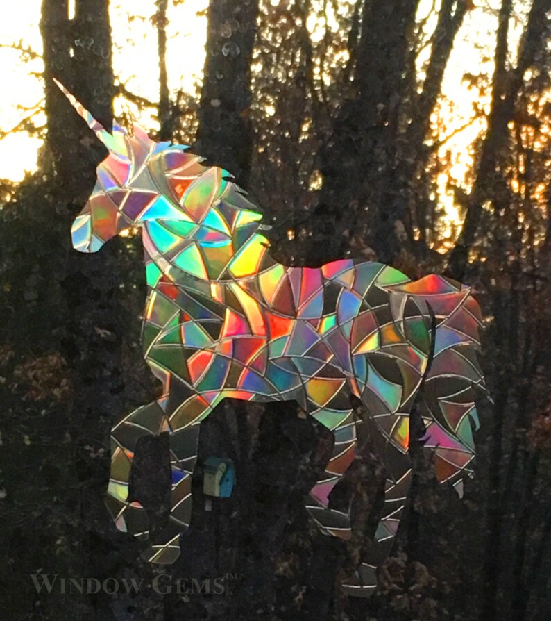 May include: A rainbow iridescent unicorn silhouette window cling. The unicorn is made of a holographic material that reflects light in a variety of colors. The unicorn is standing in front of a background of trees.