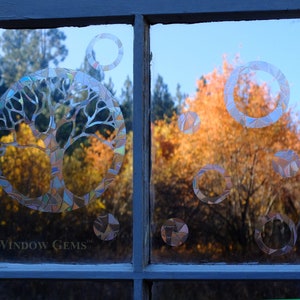 May include: A window with a decorative tree of life design and circles on the glass. The tree is made of iridescent mosaic pieces. The background is a blurry view of fall foliage.