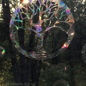 May include: A iridescent window cling featuring a tree design with a circular shape. The cling is made of a holographic material that reflects light in a rainbow of colors. The text "WINDOW GEMS&trade;" is visible in the bottom left corner of the image.