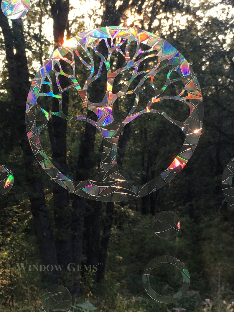 May include: A iridescent window cling featuring a tree design with a circular shape. The cling is made of a holographic material that reflects light in a rainbow of colors. The text "WINDOW GEMS&trade;" is visible in the bottom left corner of the image.