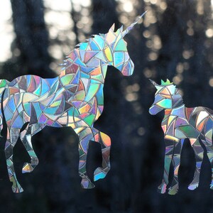 May include: Two iridescent unicorn silhouettes, one large and one small, are cut from a reflective material. The unicorns are facing the right side of the image. The background is a blurry image of trees.