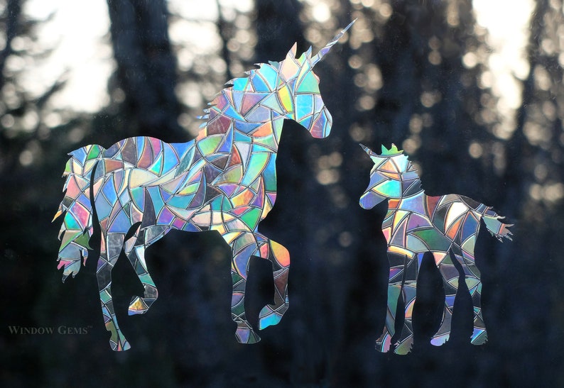 May include: Two iridescent unicorn silhouettes, one large and one small, are cut from a reflective material. The unicorns are facing the right side of the image. The background is a blurry image of trees.