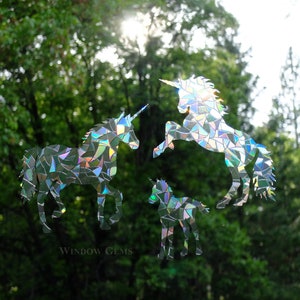 May include: Three iridescent unicorn silhouettes made from mosaic pieces of recycled CDs. The unicorns are in various poses, with one standing, one rearing up, and one standing with its head lowered. The background is a blurry image of green trees.