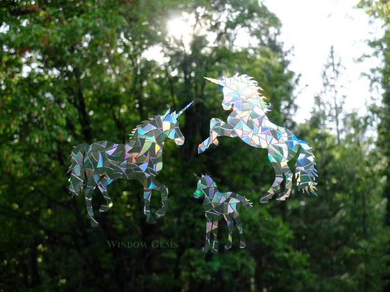 May include: Three iridescent unicorn silhouettes made from mosaic pieces of recycled CDs. The unicorns are in various poses, with one standing, one rearing up, and one standing with its head lowered. The background is a blurry image of green trees.