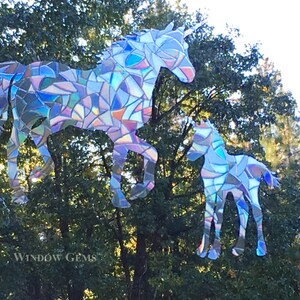 May include: A pair of iridescent unicorn silhouettes made from mosaic glass pieces. The larger unicorn is standing with its head turned to the side, while the smaller unicorn is standing with its head facing forward. The unicorns are set against a backdrop of green trees.