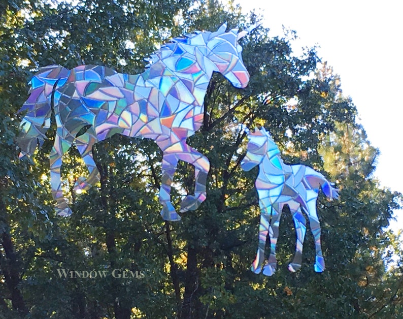 May include: A pair of iridescent unicorn silhouettes made from mosaic glass pieces. The larger unicorn is standing with its head turned to the side, while the smaller unicorn is standing with its head facing forward. The unicorns are set against a backdrop of green trees.
