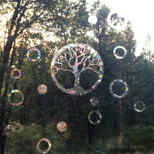 May include: A colorful iridescent window cling featuring a tree of life design with a circle of smaller circles around it. The cling is placed on a window with a view of trees and grass. The text "WINDOW GEMS&trade;" is visible in the bottom right corner.