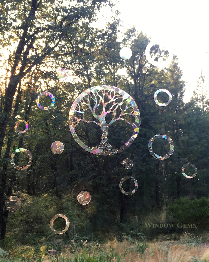 May include: A colorful iridescent window cling featuring a tree of life design with a circle of smaller circles around it. The cling is placed on a window with a view of trees and grass. The text "WINDOW GEMS&trade;" is visible in the bottom right corner.