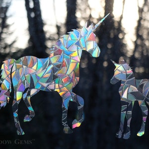 May include: Two iridescent unicorn window clings, one large and one small, with a mosaic-like design. The larger unicorn is facing left, while the smaller unicorn is facing right. The background is a blurred image of trees.