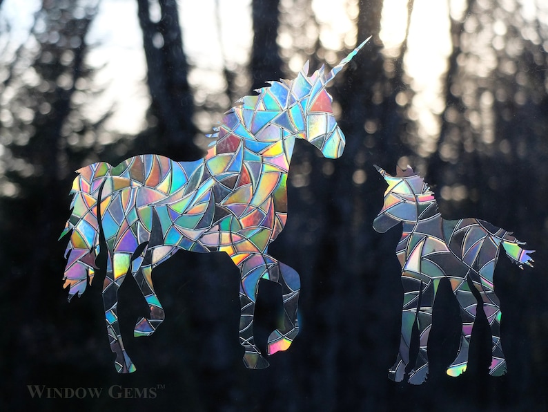 May include: Two iridescent unicorn window clings, one large and one small, with a mosaic-like design. The larger unicorn is facing left, while the smaller unicorn is facing right. The background is a blurred image of trees.