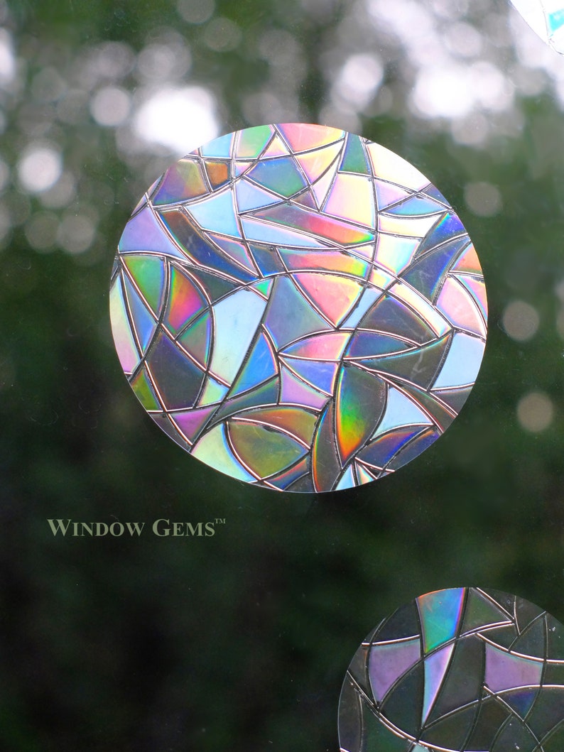 May include: Two iridescent window clings in the shape of circles. The larger circle has a geometric design with a mix of blue, pink, and purple hues. The smaller circle has a similar design with black, purple, and blue hues. The text "WINDOW GEMS&trade;" is visible at the bottom of the image.