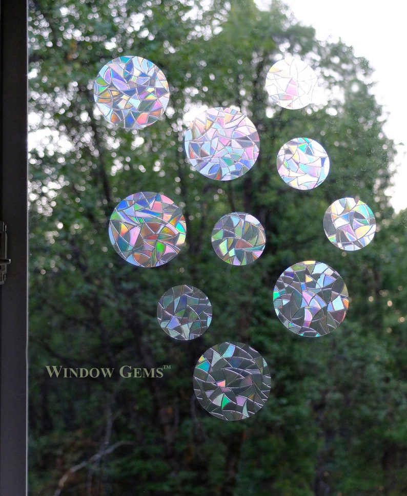 May include: A set of iridescent window clings in the shape of circles. The clings are made of a clear material with a rainbow-like pattern. The clings are arranged in a random pattern on a window. The text "WINDOW GEMS&trade;" is visible in the bottom left corner of the image.