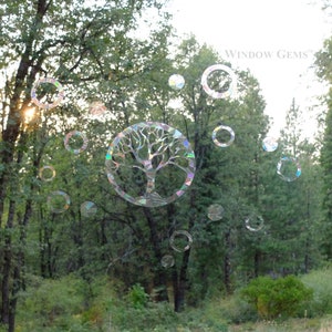 May include: A window with a tree of life sticker and iridescent circles on it. The window is looking out onto a forest with green trees and bushes. The text "WINDOW GEMS" is visible in the top right corner of the image.