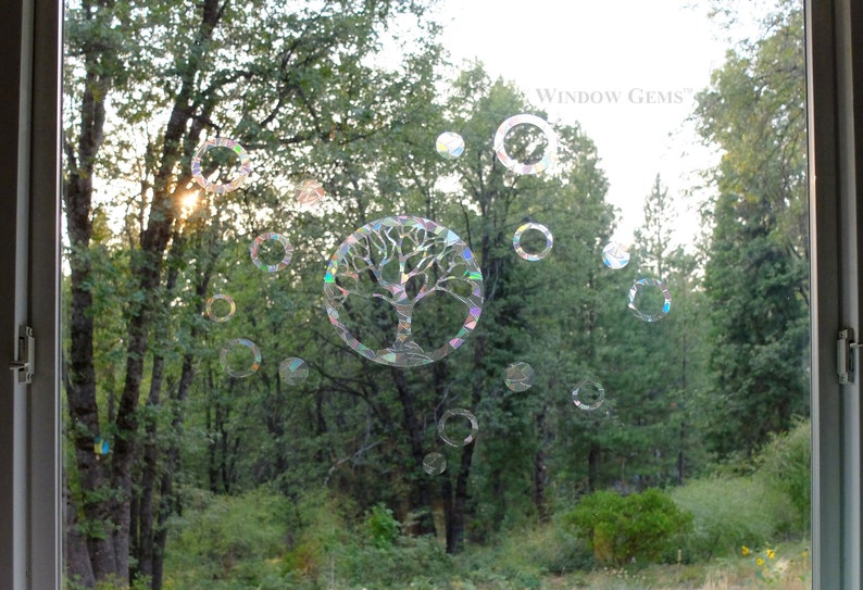 May include: A window with a tree of life sticker and iridescent circles on it. The window is looking out onto a forest with green trees and bushes. The text "WINDOW GEMS" is visible in the top right corner of the image.