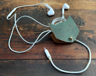May include: A green leather cord organiser shaped like a cat's head with white headphone cords wrapped around it.