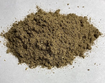 May include: A pile of finely ground, brownish-green powder, possibly a spice or herb blend, sits on a white surface. The texture appears granular with visible plant fibers. The lighting is soft, highlighting the natural color and texture of the powder.