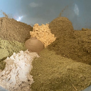 Op de afbeelding: Een lichtblauwe kom gevuld met diverse gedroogde kruiden en specerijen. Het assortiment omvat tinten groen, bruin en beige poeders en korrels, wat duidt op een culinaire of kruidenbereiding.