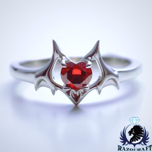 May include: A silver ring with a heart-shaped, deep red gemstone. The ring's design incorporates stylized, pointed elements around the gem, creating a unique and elegant appearance. The ring is set against a white background.