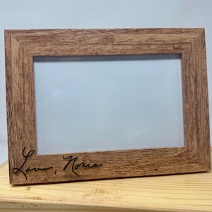 May include: A rectangular picture frame with a light brown wood grain finish. The frame features a white insert and the black cursive text "Love, Nonie" engraved on the bottom left corner of the wooden border, creating a clear and simple display.