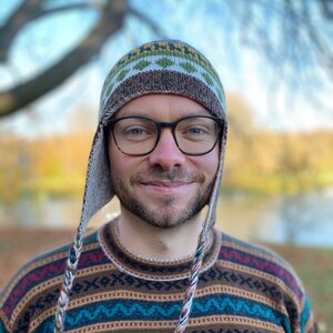 May include: A knitted hat with ear flaps and a matching sweater. The hat features a geometric pattern in shades of green, brown, and white. The sweater has a similar pattern in brown, blue, red, and white. The ear flaps have braided tassels.