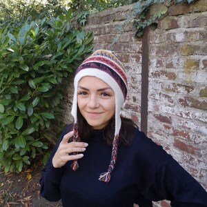 May include: A white, red, and blue patterned knit hat with ear flaps and braided tassels. The hat has a colorful geometric design and is worn by a person. The hat is a winter accessory.