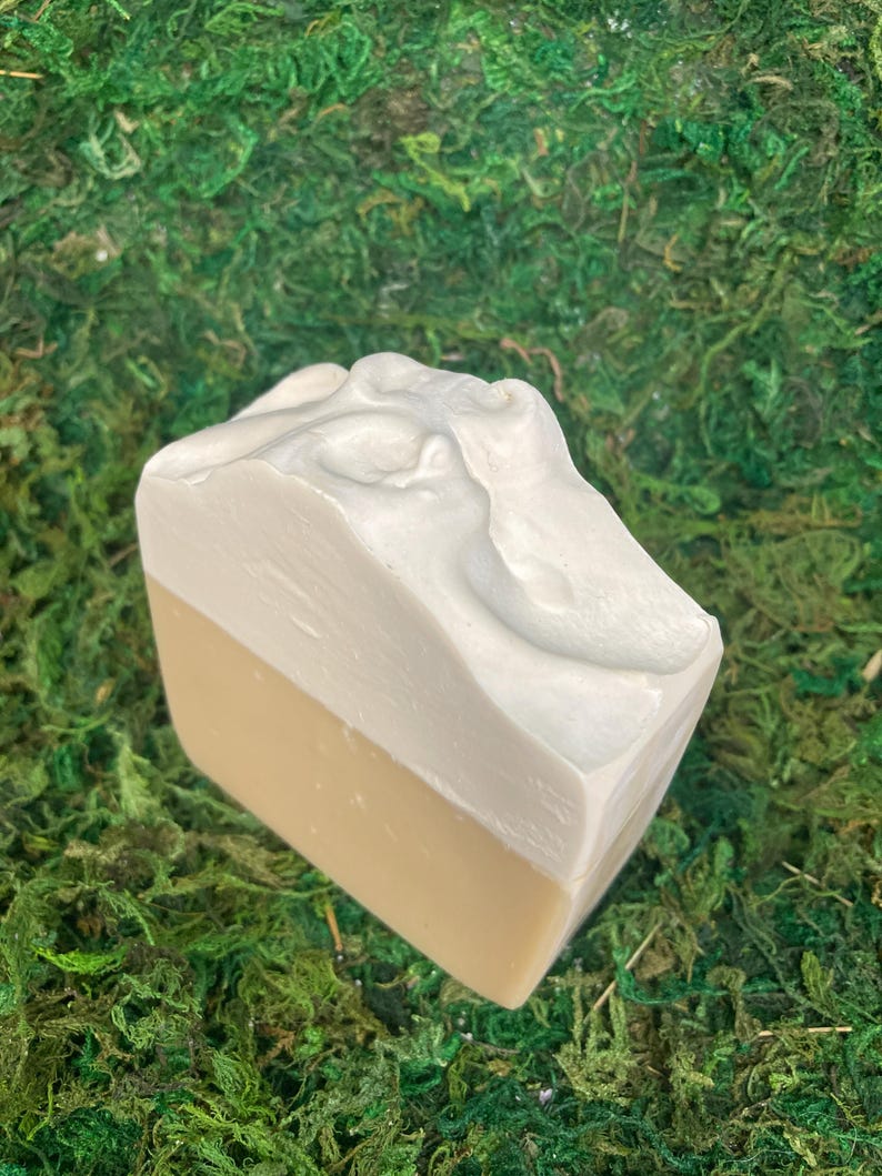 May include: A two-toned bar of soap with a light beige bottom and a white, textured top. The soap is rectangular and sits on a bed of green moss. The top of the soap has a sculpted design.