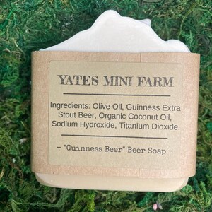 May include: A bar of "Guinness Beer" soap from Yates Mini Farm. The soap is wrapped in a brown paper label with the ingredients listed. The soap has a light beige color with a white, foamy top. The background is green.