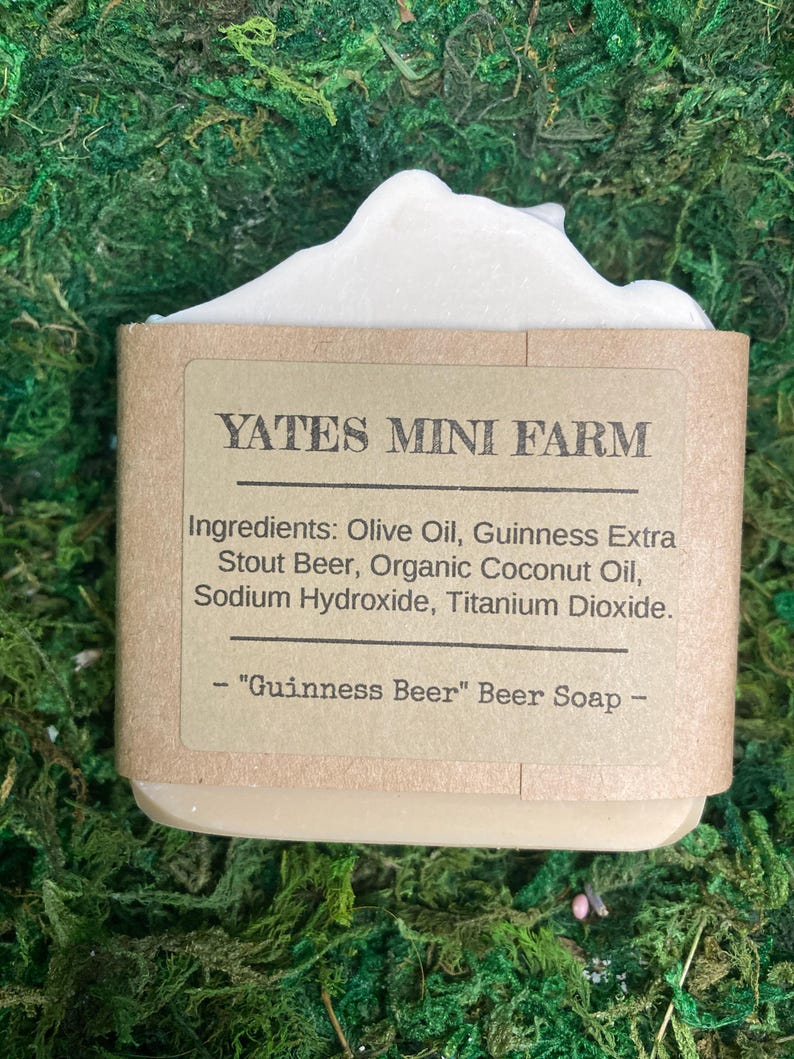 May include: A bar of "Guinness Beer" soap from Yates Mini Farm. The soap is wrapped in a brown paper label with the ingredients listed. The soap has a light beige color with a white, foamy top. The background is green.