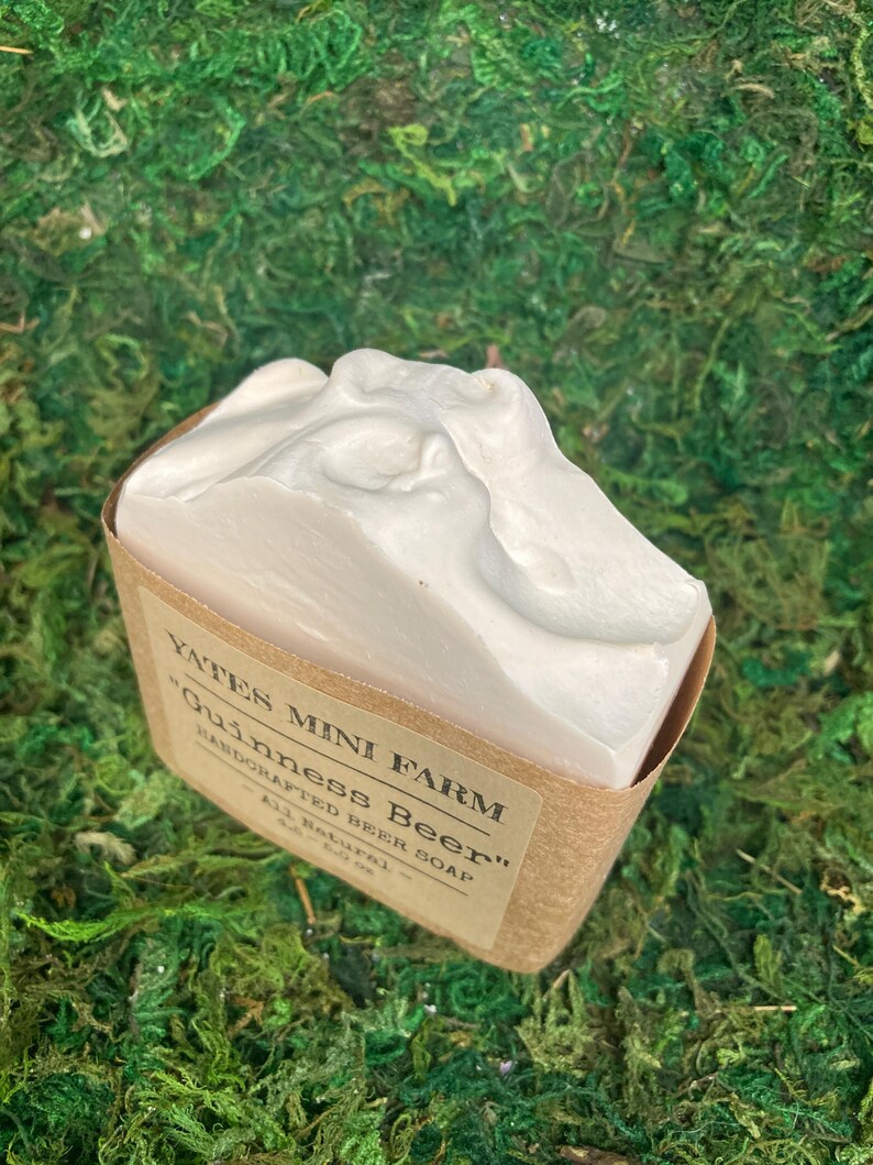 May include: A white bar of "Guinness Beer" soap from Yates Mini Farm, in a brown paper wrapper. The soap has a textured, foamy top. The label reads "Handcrafted Beer Soap - All Natural - 4.0 oz."