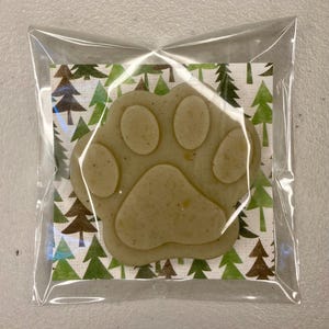 May include: A light tan paw-shaped item, possibly a treat or soap, inside a clear plastic bag. The paw print is on a patterned card with green and brown Christmas trees. The background is a light color.