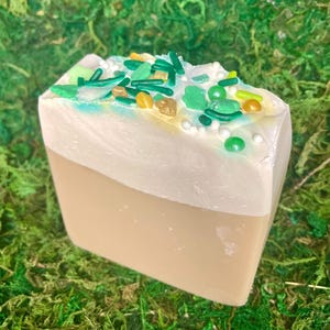 May include: A bar of soap resembling a slice of cake, with a light brown base and a white top decorated with green, yellow, and gold sprinkles. The soap is set against a backdrop of green moss.