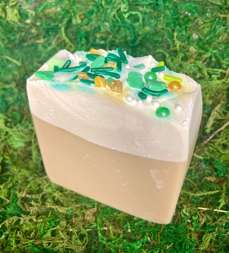 May include: A bar of soap resembling a slice of cake, with a light brown base and a white top decorated with green, yellow, and gold sprinkles. The soap is set against a backdrop of green moss.
