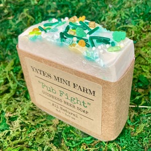 May include: A bar of "Pub Fight" Guinness beer soap from Yates Mini Farm, decorated with green, yellow, and white sprinkles. The soap is wrapped in a brown paper label and sits on a bed of green moss.