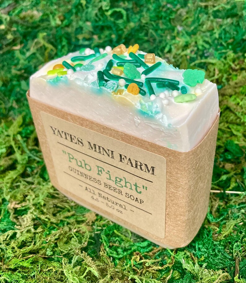 May include: A bar of "Pub Fight" Guinness beer soap from Yates Mini Farm, decorated with green, yellow, and white sprinkles. The soap is wrapped in a brown paper label and sits on a bed of green moss.