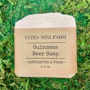 May include: A bar of Guinness Beer Soap from Yates Mini Farm. The soap is light beige with a brown paper label. The label reads "YATES MINI FARM", "Guinness Beer Soap", and "- HANDCRAFTED & VEGAN - 4-5 oz". The soap is on a bed of green moss.