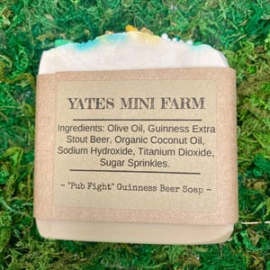 May include: A bar of soap with a light-colored base, topped with blue, yellow, and green sprinkles. The label reads "YATES MINI FARM" and lists ingredients including Guinness Stout Beer. The soap is labeled "Pub Fight" Guinness Beer Soap.