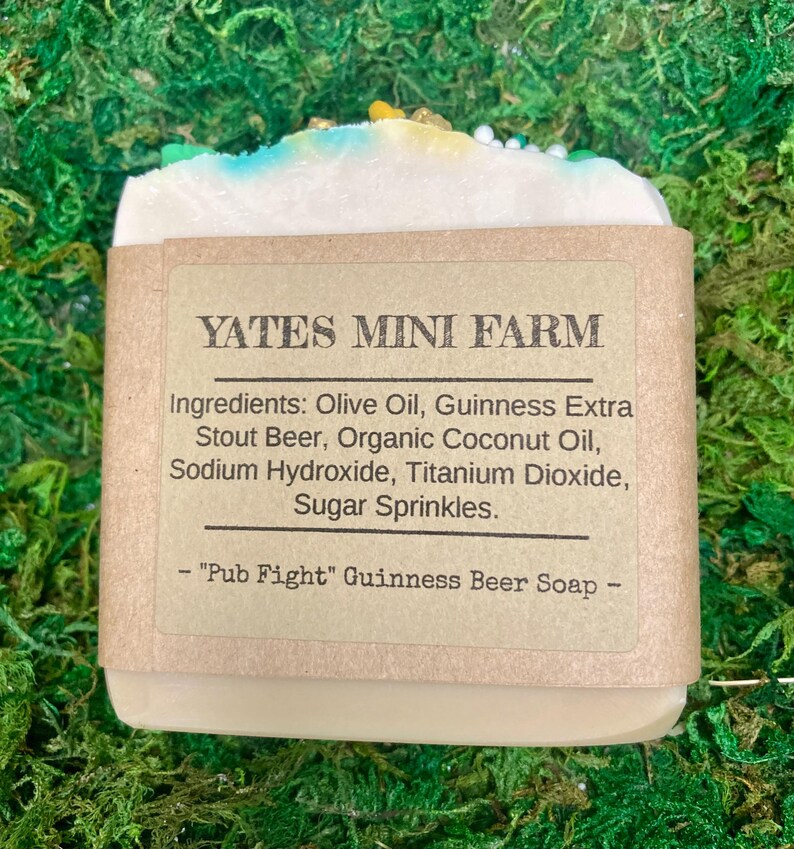 May include: A bar of soap with a light-colored base, topped with blue, yellow, and green sprinkles. The label reads "YATES MINI FARM" and lists ingredients including Guinness Stout Beer. The soap is labeled "Pub Fight" Guinness Beer Soap.