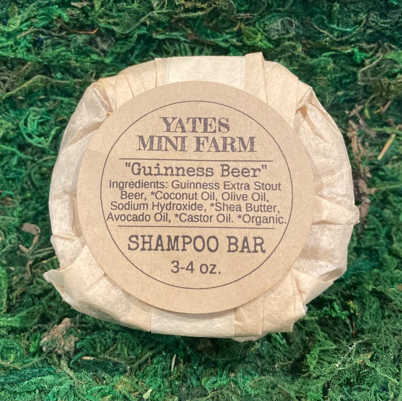 Op de afbeelding: Een bruine, in papier gewikkelde shampoo-bar met het label "Yates Mini Farm" en de tekst "Guinness Beer" en een lijst met ingredi&euml;nten, waaronder Guinness Extra Stout bier, kokosolie, olijfolie, natriumhydroxide, sheaboter, avocado-olie, ricinusolie en biologische ingredi&euml;nten. De bar weegt 3-4 ounce.