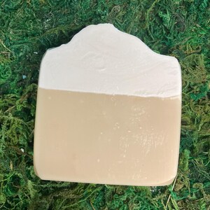 May include: A rectangular bar of soap with a two-tone design. The top half is a creamy white, and the bottom half is a light tan color. The soap has a slightly uneven, textured surface and is set against a backdrop of green moss.