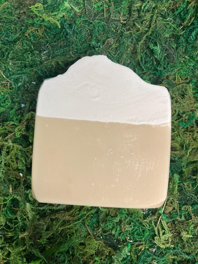 May include: A rectangular bar of soap with a two-tone design. The top half is a creamy white, and the bottom half is a light tan color. The soap has a slightly uneven, textured surface and is set against a backdrop of green moss.