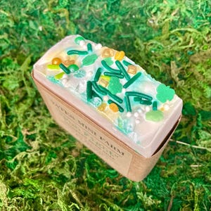 May include: A rectangular bar of soap with a white top decorated with green sprinkles, shamrocks, and gold accents. The soap sits in a brown paper box, and the background is green moss. The soap is labeled "Yates Mini Farm".