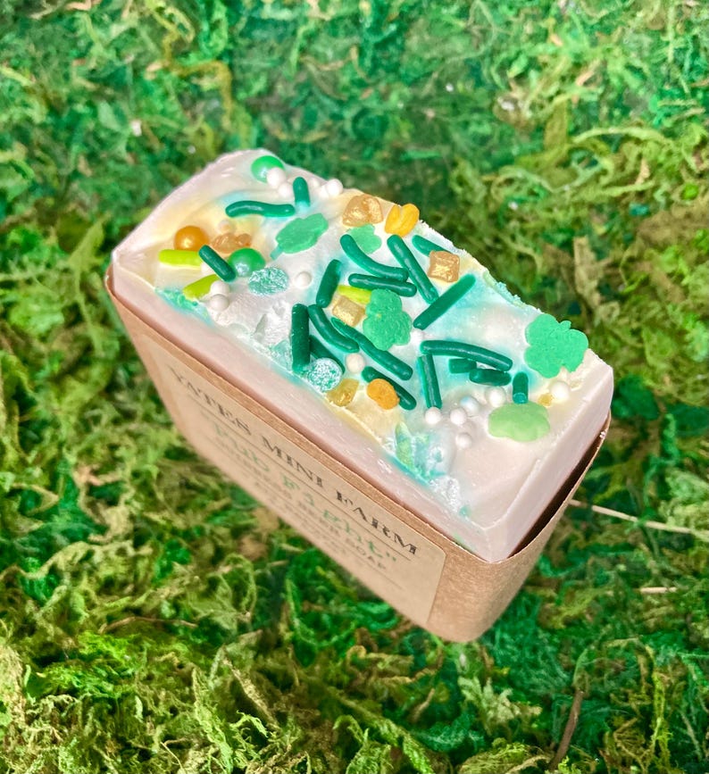 May include: A rectangular bar of soap with a white top decorated with green sprinkles, shamrocks, and gold accents. The soap sits in a brown paper box, and the background is green moss. The soap is labeled "Yates Mini Farm".