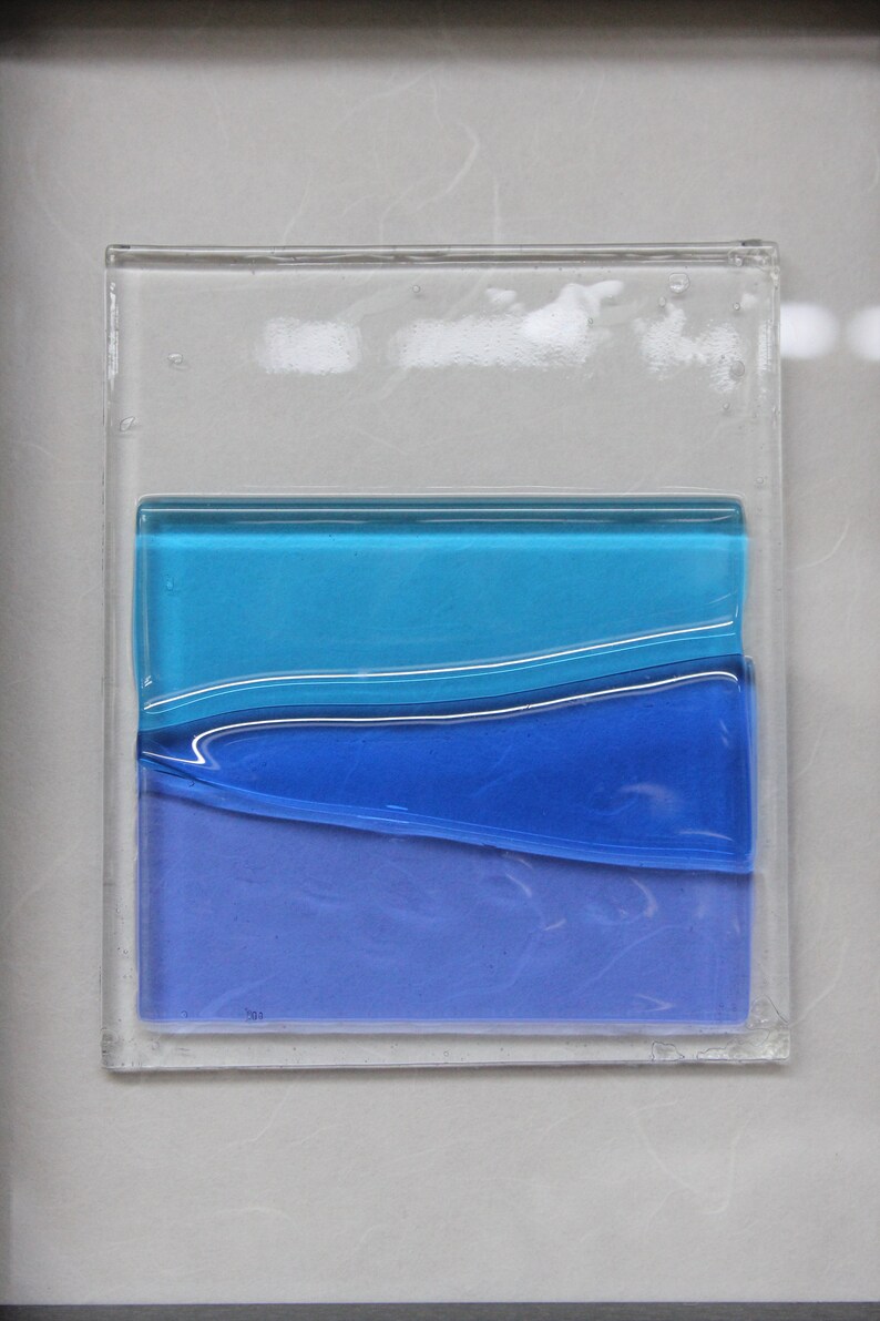 May include: A framed piece of art with layered blue glass. The glass is layered in shades of blue, from light to dark, and the layers are slightly wavy.