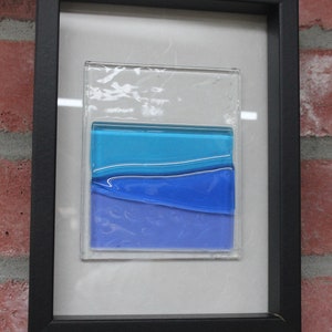 May include: A framed piece of art featuring a layered glass design with blue and purple hues. The glass is set against a white background and framed in black.