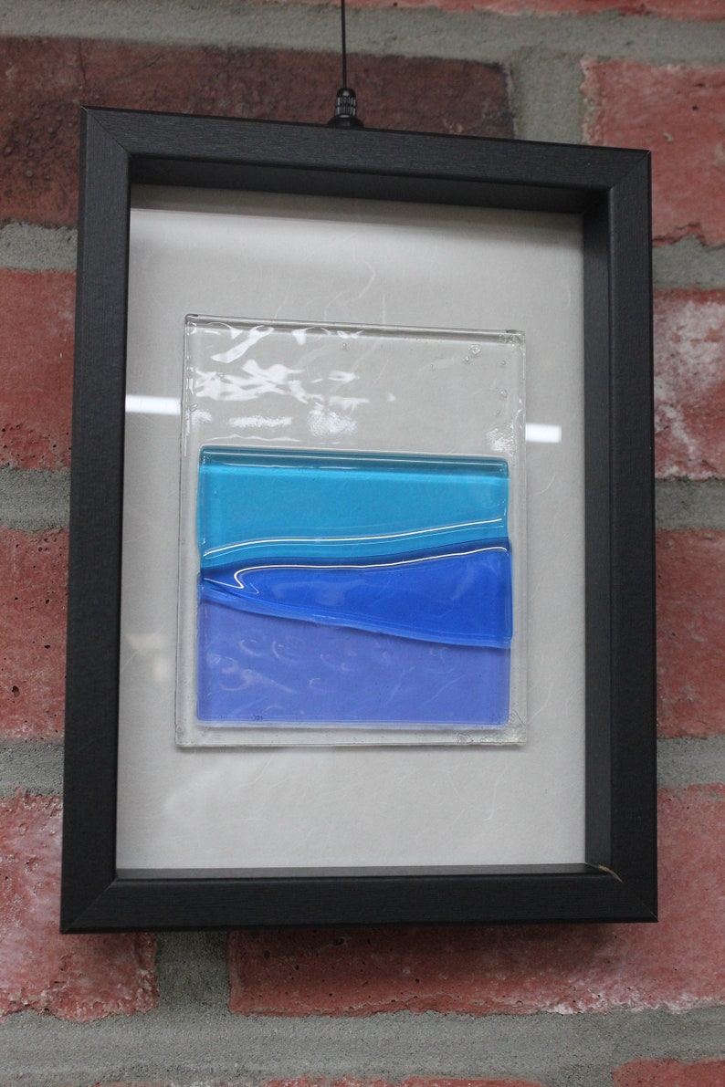 May include: A framed piece of art featuring a layered glass design with blue and purple hues. The glass is set against a white background and framed in black.