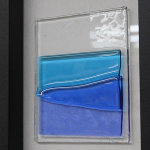 May include: A framed piece of art featuring layered glass panels in shades of blue. The glass panels are arranged in a wave-like pattern, creating a sense of depth and movement.