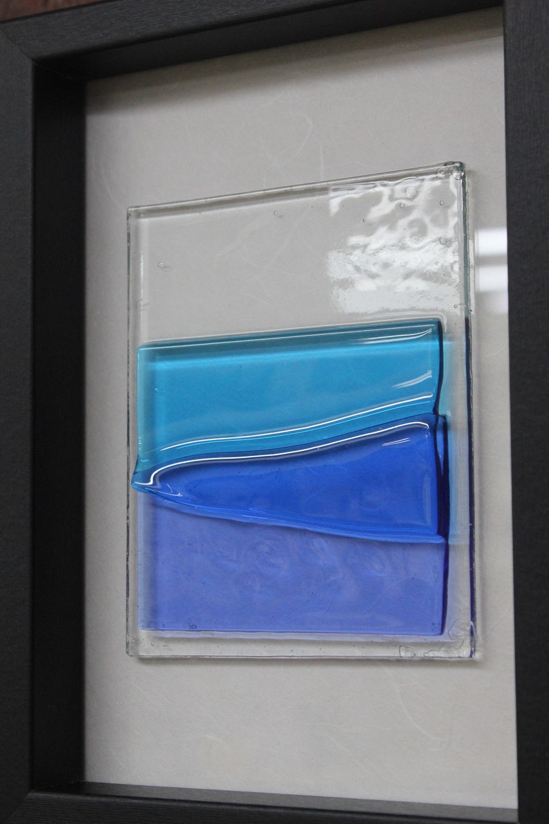 May include: A framed piece of art featuring layered glass panels in shades of blue. The glass panels are arranged in a wave-like pattern, creating a sense of depth and movement.